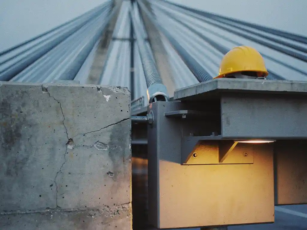 Stalen kabelbrug verbindt verweerd betonnen platform met versterkt platform, gele helm op stabiele zijde, amberkleurig licht, lage camerahoek.